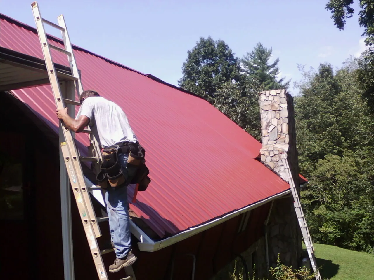 Roofer on a ladder beside a red metal roof and stone chimney — Soffit & Fascia Repair by Roofing Brothers in Oconomowoc